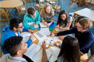 Diverse group of teen high school students are sitting around round table in library.
